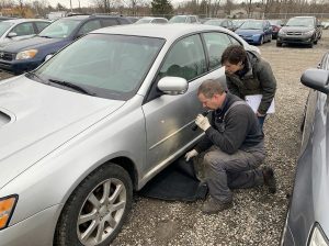 This image shows a used car buyer inspecting a vehicle for flood damage, supporting a guide about musty smells, damp carpets, rust, electronics, title history, VIN checks, and pre-purchase inspections.
